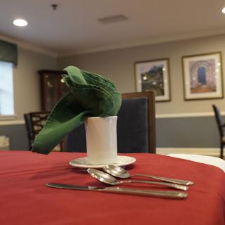 A dining table set with a green napkin in a cup and cutlery arranged neatly on a red tablecloth.
