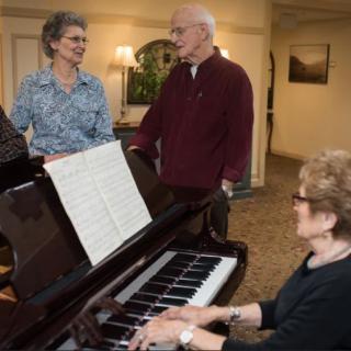 A group of elderly people enjoying music around a piano indoors.