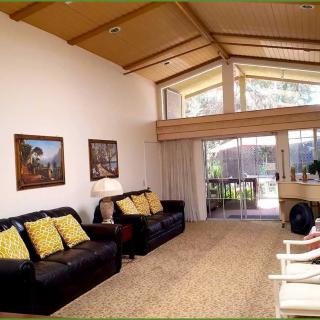 A spacious living room with a high ceiling, featuring comfortable sofas, decorative artwork on the walls, and large windows allowing natural light to fill the space.