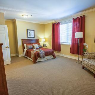 A cozy bedroom with a bed, chair, and table lamp, featuring red curtains and warm lighting.
