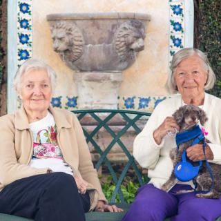 Two elderly women sitting on a bench, one holding a small dog, and both smiling.