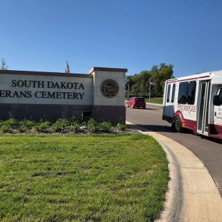 A bus approaches the entrance sign of South Dakota Veterans Cemetery on a sunny day.