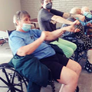 A group of individuals in wheelchairs participate in a seated exercise session while wearing masks.