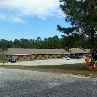 A one-story building with a parking lot, surrounded by trees under a partly cloudy sky.