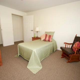 A minimalist bedroom with a single bed, green bedding, a nightstand with a lamp, and a rocking chair on a carpeted floor.