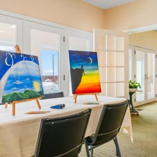 A bright room set up for an art class with paintings on easels and seating arranged around tables.