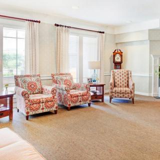 A bright living room featuring floral armchairs, large windows with curtains, and a classic clock.