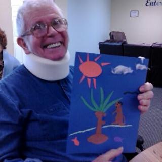 An elderly person wearing a neck brace smiles while holding up a colorful painting of a sunny landscape.