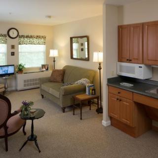 A cozy living area with a sofa, kitchenette, and computer desk, well-lit by natural light from the window.