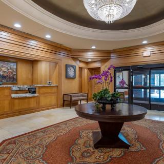 A well-lit hotel lobby featuring a chandelier, a round table with a floral arrangement, and elegant wood paneling.