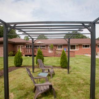A view of a garden featuring a pergola and rustic chairs, set against a backdrop of brick buildings.