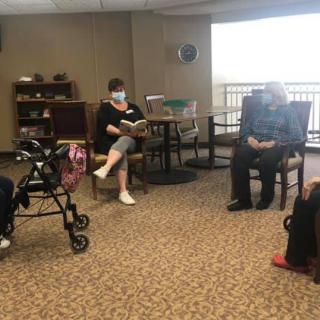 A group of people gathered indoors, with one person reading to the others in a community setting.