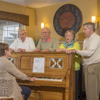 A group of elderly people gathered around a piano, enjoying a singing session indoors.