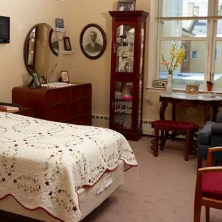 A cozy vintage-style bedroom with classic furniture, including a bed with a lace cover, a dresser with a mirror, and a small table by the window.