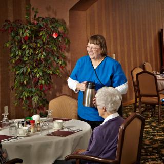 A caregiver serving a meal to an elderly woman in a dining room setting.