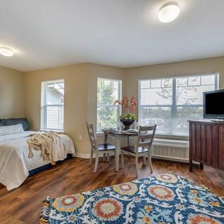 A cozy studio apartment featuring a bed, dining table, and television, with large windows and hardwood flooring.