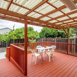 A cozy outdoor patio with a wooden deck and pergola, featuring a set of chairs and a table, surrounded by greenery.