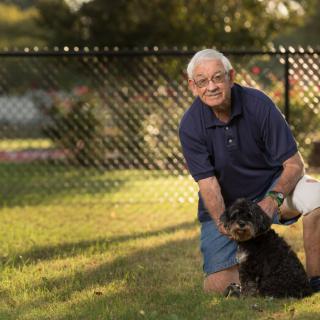 An elderly man kneeling on grass next to a dog in a sunny yard.