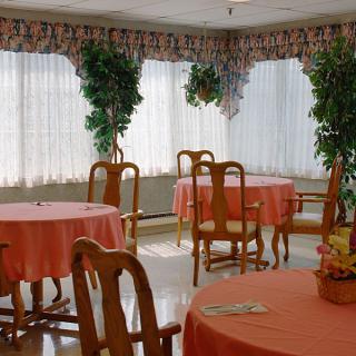 A cozy dining area with round tables covered in coral tablecloths, surrounded by wooden chairs and adorned with potted plants near the windows.