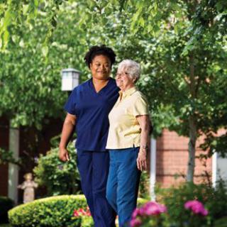 A caregiver walking with an elderly woman in a garden setting.