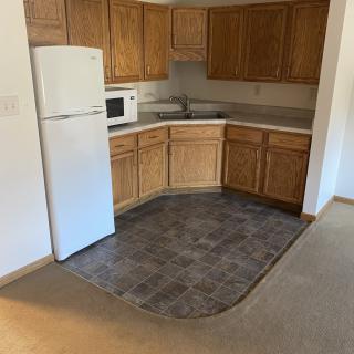 An empty apartment kitchen with wooden cabinets, a white fridge, and a microwave.