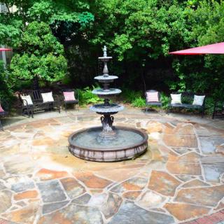 A tranquil outdoor patio with a central multi-tiered fountain surrounded by stone flooring and chairs, shaded by greenery.