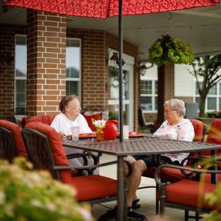 Two elderly women enjoying a conversation under a red umbrella on a patio.