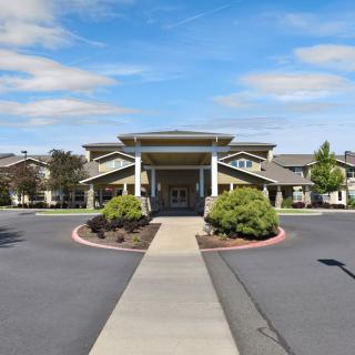 A wide pathway leads to the entrance of a large building, surrounded by neatly trimmed shrubs and landscaping under a partly cloudy sky.