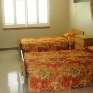 A simple hospital room featuring two twin beds with orange floral bedding, a window with curtains, and minimal furnishings.
