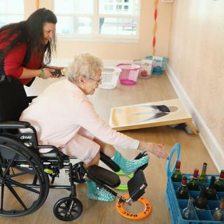 An elderly woman in a wheelchair is engaged in an art therapy session, assisted by a caregiver.