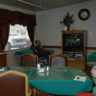 A person sitting in a room with a television, chairs, and a table.