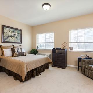 A cozy bedroom with beige walls, featuring a bed, carpet flooring, and two windows allowing natural light.