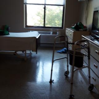 A well-lit hospital room featuring a bed, a walker, and a view outside the window.