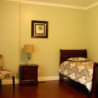 A minimalist bedroom with a single bed, nightstand, lamp, and chair on hardwood floors with green walls.