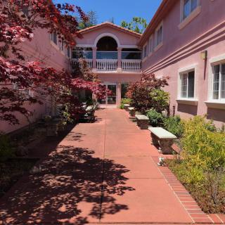 A sunny courtyard with a pathway flanked by a pink building, greenery, and benches.
