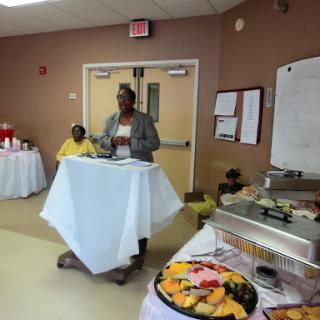 A speaker addresses an audience at a community event, with a buffet table in the foreground.
