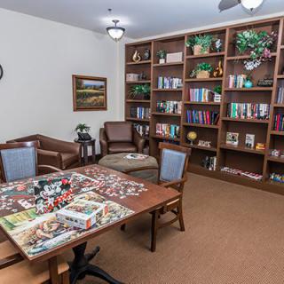 A cozy library room with bookshelves, a table partially covered with a puzzle, and comfortable seating.