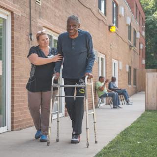 A caregiver assists an elderly man using a walker in a community setting.