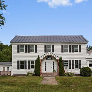 A two-story colonial-style house with a white facade and green trim, surrounded by greenery.