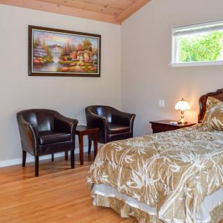 A cozy bedroom with elegant furniture and decor, featuring a bed, two chairs, a lamp, and a painting against a hardwood floor.