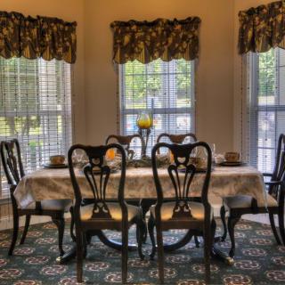A cozy dining room with a set table and elegant chairs near large windows with floral curtains.
