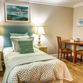 A cozy bedroom with a single bed, decorated with pillows and art, featuring a lamp and small dining table.