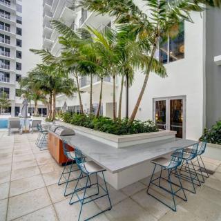 An urban outdoor patio area with tables, chairs, and palm trees surrounded by modern buildings.