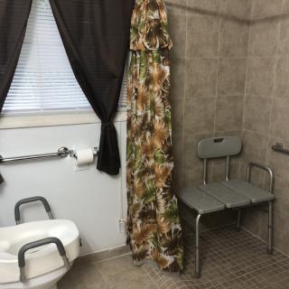 An accessible bathroom featuring a shower chair and grab bars alongside a toilet and window.