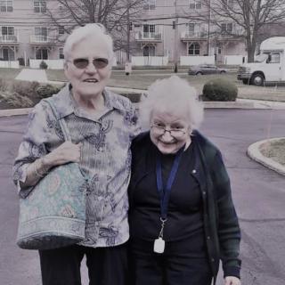 Two elderly women smiling and standing outside together.