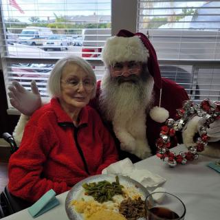 An elderly woman sits with a person dressed as Santa Claus during a festive holiday meal.