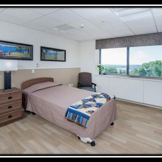 A hospital room with a single bed, nightstand, and chair, featuring large windows and framed artwork on the walls.