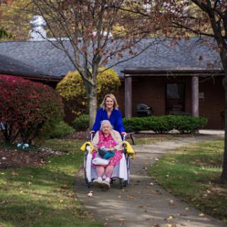 A caregiver pushes an elderly person in a wheelchair through a peaceful residential setting during autumn.