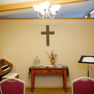 A small chapel room featuring a cross, piano, flowers on a table, and a lectern.