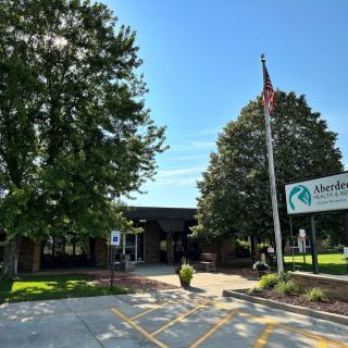 A building entrance surrounded by trees with an American flag and a sign on a sunny day.